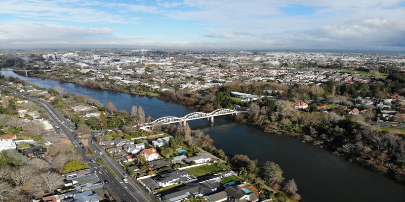 Aerial view of Hamilton, New Zealand, with a river, white bridge, tree-lined neighborhoods, and a main road. A sprawling urban landscape stretches under a partly cloudy sky.