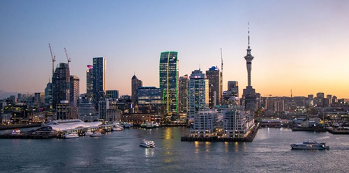 Skyline of Auckland, New Zealand, at dusk with the iconic Sky Tower and high-rise buildings illuminated against a clear evening sky. Boats and ferries move through the harbor in the foreground, reflecting city lights on the water.