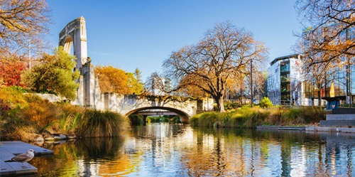 Skyline of Christchurch, New Zealand, with buildings along a waterway and a bridge visible in the distance. The scene includes modern architecture, trees, and reflections in the calm water, capturing the city’s urban landscape.