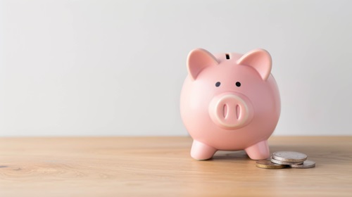 A pink piggy bank sitting on a wooden surface next to a small stack of coins, symbolising cost savings and financial efficiency. The image represents the potential savings achieved through energy-efficient air conditioning and heat pump solutions.