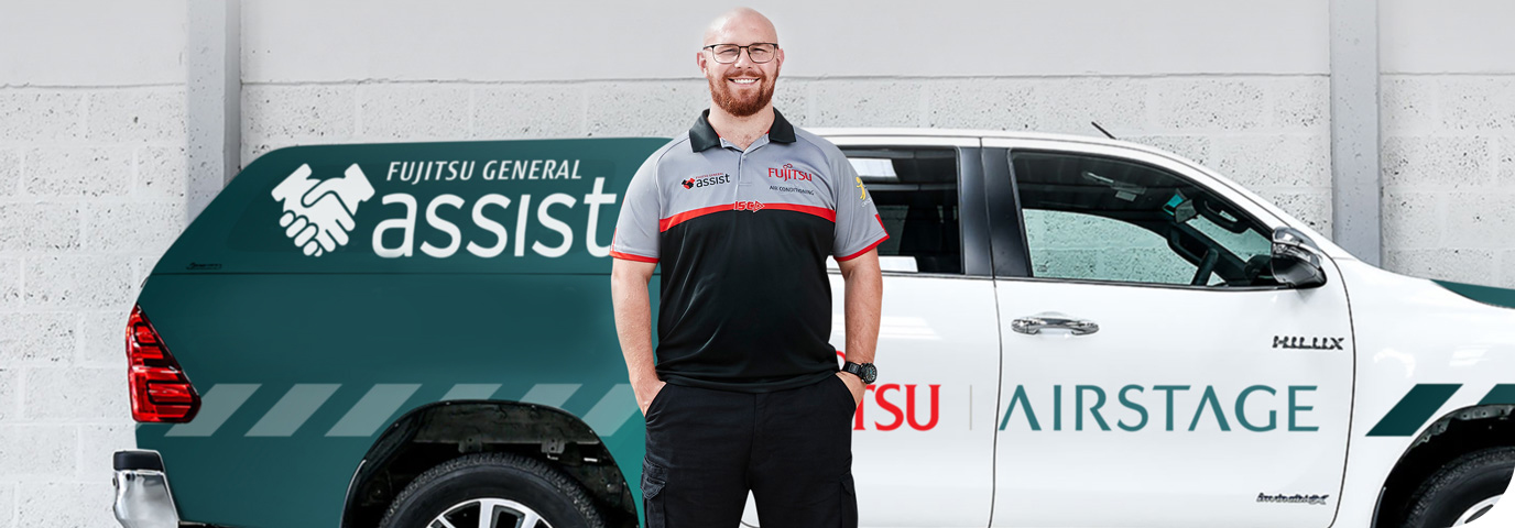 A smiling heat pump & air conditioner technician stands in front of a Fujitsu General Assist service vehicle. Wearing a branded Fujitsu uniform, the white vehicle displays "Fujitsu Airstage" and "Fujitsu General Assist" logos, plus a handshake graphic.